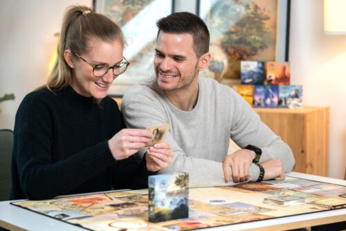 "Boîte de jeu CARTAVENTURA : COSMOLOGIA posée sur une table en bois, entourée de cartes illustrées représentant des scènes historiques et fantastiques."