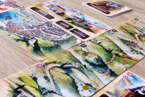 "Boîte de jeu CARTAVENTURA : COSMOLOGIA posée sur une table en bois, entourée de cartes illustrées représentant des scènes historiques et fantastiques."